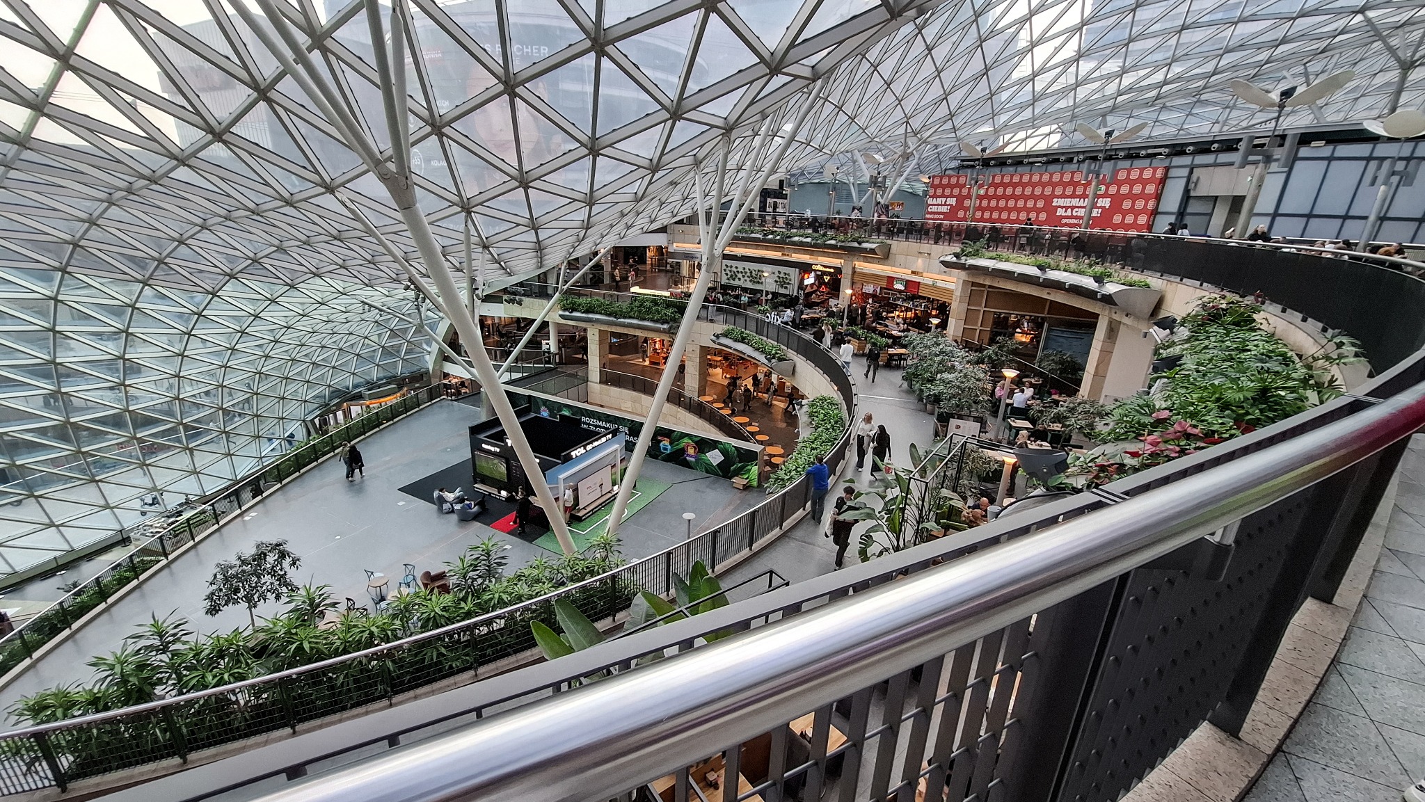 Wnętrze nowoczesnego centrum handlowego z przeszklonym dachem, widok z góry na kilka poziomów z roślinnością, liczne lokale gastronomiczne i stoiska reklamowe.