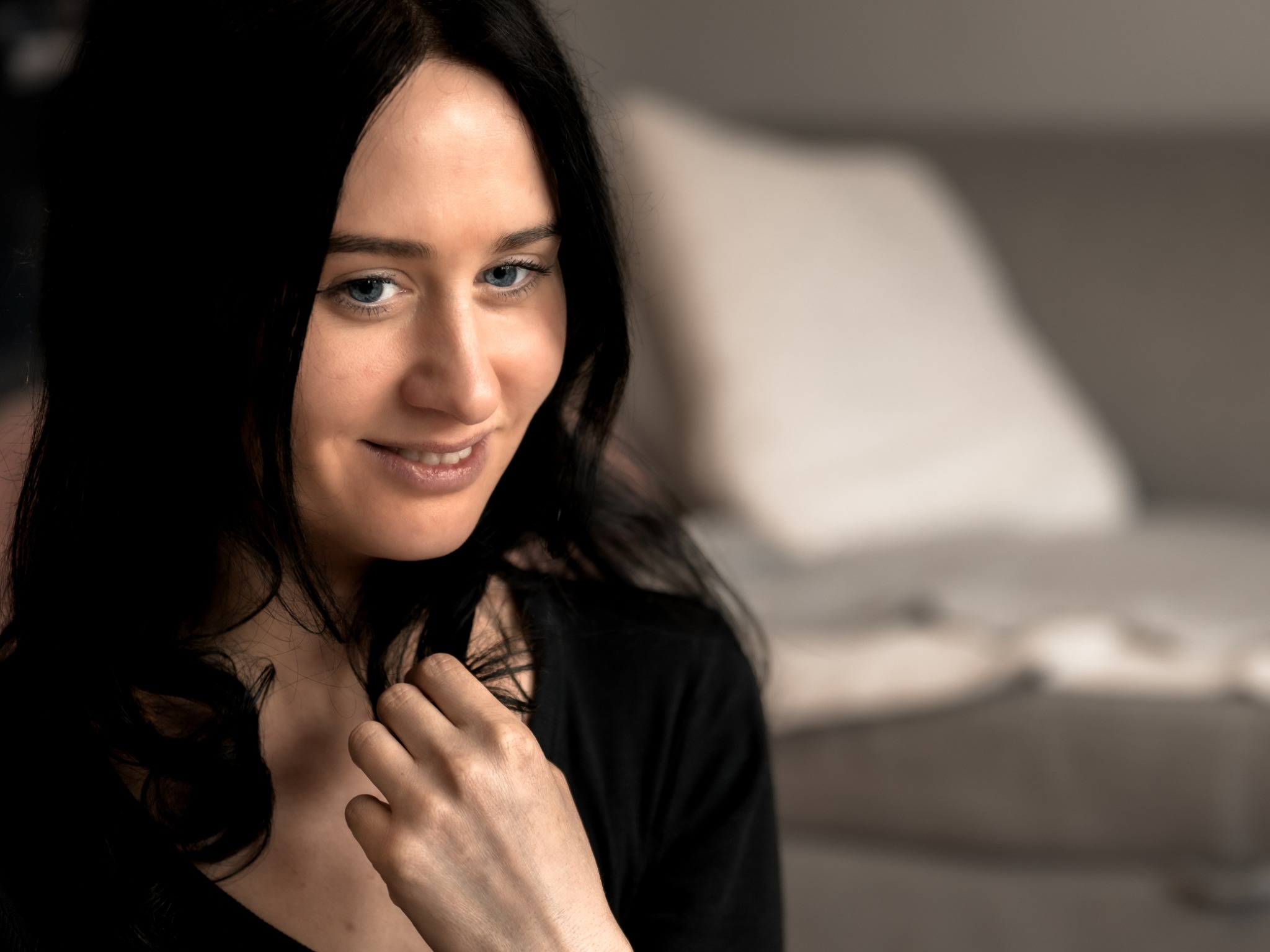 Portret uśmiechniętej kobiety o niebieskich oczach i czarnych włosach, delikatnie dotykającej włosów, z rozmytym tłem wnętrza.