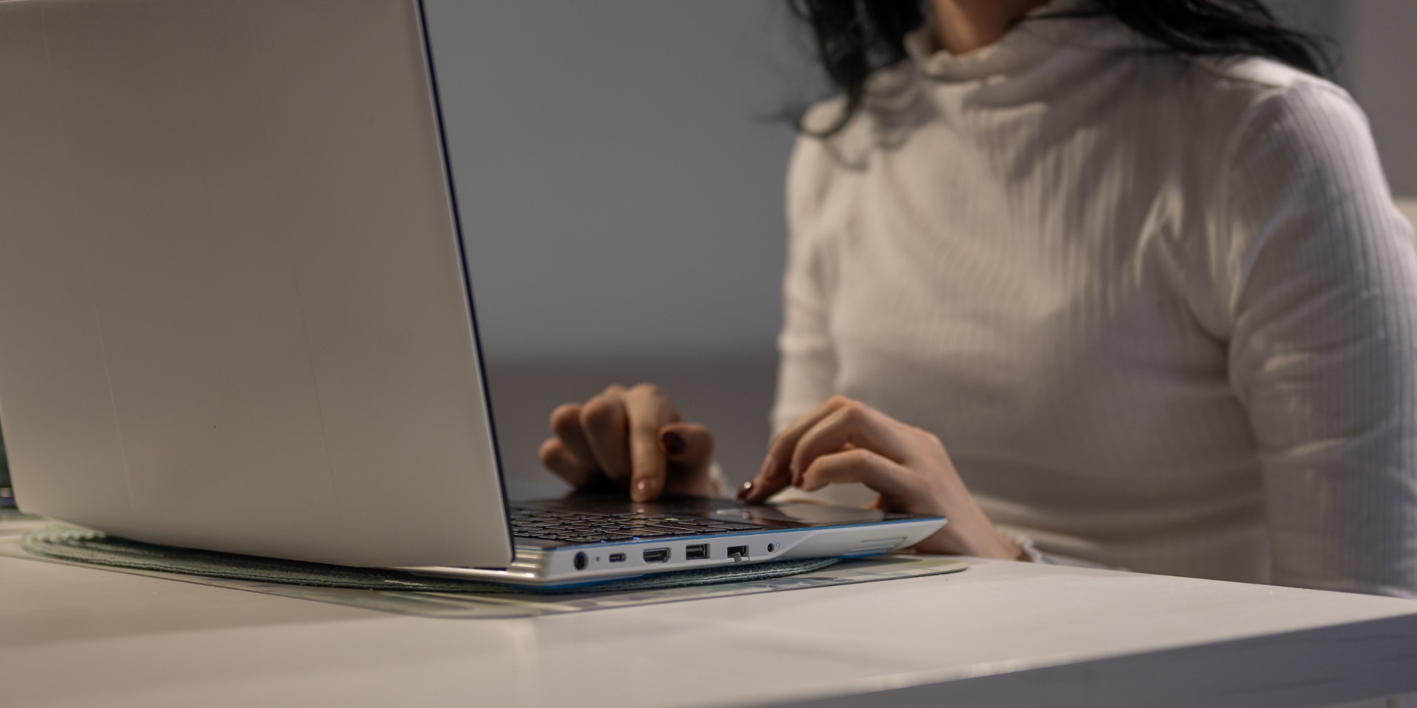 Kobieta w białej bluzce pisze na laptopie z niebieskimi akcentami, umieszczonym na zielonej podkładce na białym stole.