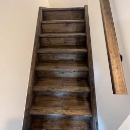 MODERN THERMO BUILDINGS SPÓŁKA Z OGRANICZONĄ ODPOWIEDZIALNOŚCIĄ - Schody drewniane z ciemnego drewna, widok od dołu, z widoczną poręczą na ścianie pomalowanej na biało, oświetlenie górne.