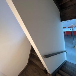 MODERN THERMO BUILDINGS SPÓŁKA Z OGRANICZONĄ ODPOWIEDZIALNOŚCIĄ - Nowoczesne schody drewniane z gładkimi, jasnymi ścianami i minimalistyczną balustradą, widok na jadalnię z czerwonymi krzesłami w tle.