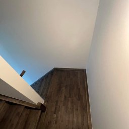 MODERN THERMO BUILDINGS SPÓŁKA Z OGRANICZONĄ ODPOWIEDZIALNOŚCIĄ - Widok z dołu na drewniane schody z białą balustradą, prowadzące w górę, z widocznym fragmentem ściany i sufitu.