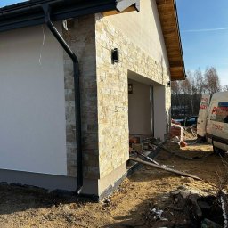 MODERN THERMO BUILDINGS SPÓŁKA Z OGRANICZONĄ ODPOWIEDZIALNOŚCIĄ - Fragment elewacji domu jednorodzinnego z wykończeniem z kamienia dekoracyjnego, widoczny czarny system rynnowy i fragment ocieplonej ściany, w tle zaparkowany samochód dostawczy ekipy budowlanej.