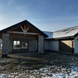 MODERN THERMO BUILDINGS SPÓŁKA Z OGRANICZONĄ ODPOWIEDZIALNOŚCIĄ - Nowoczesny dom jednorodzinny w trakcie wykończenia elewacji, z widocznym fragmentem otynkowanej części i kamienną kolumnadą przy wejściu, otoczony ziemią pokrytą resztkami śniegu.