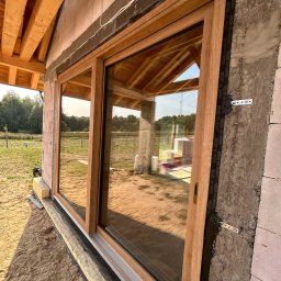 MODERN THERMO BUILDINGS SPÓŁKA Z OGRANICZONĄ ODPOWIEDZIALNOŚCIĄ - Montaż okna przesuwnego z drewnianą ramą w surowym stanie budynku z betonu komórkowego. Widoczna taśma uszczelniająca okno oraz elementy mocujące ramę. W tle widoczny krajobraz.