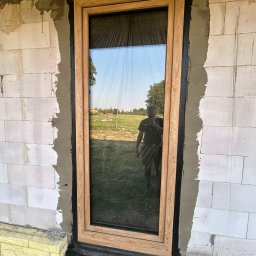 MODERN THERMO BUILDINGS SPÓŁKA Z OGRANICZONĄ ODPOWIEDZIALNOŚCIĄ - Montaż okna z drewnopodobną ramą w surowej ścianie z betonu komórkowego; widoczna izolacja, czarna uszczelka i odbicie fotografa w szybie.