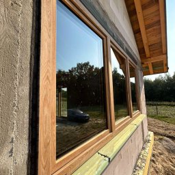 MODERN THERMO BUILDINGS SPÓŁKA Z OGRANICZONĄ ODPOWIEDZIALNOŚCIĄ - Montaż okien z drewnianą ramą w surowym budynku z betonu komórkowego; widoczny zielony materiał izolacyjny i odbicie samochodu w szybie.