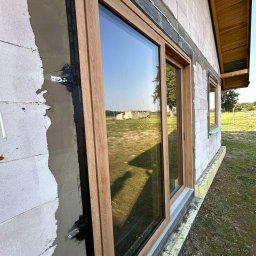 MODERN THERMO BUILDINGS SPÓŁKA Z OGRANICZONĄ ODPOWIEDZIALNOŚCIĄ - Montaż okna z drewnopodobną ramą w surowym murze z betonu komórkowego. Widoczna taśma uszczelniająca okno, ocieplenie fundamentu i odbicie krajobrazu w szybie.