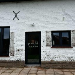 MODERN THERMO BUILDINGS SPÓŁKA Z OGRANICZONĄ ODPOWIEDZIALNOŚCIĄ - Biała, ceglana ściana budynku z zamontowanymi czarnymi oknami PCV i drzwiami. Na ścianie widoczne dekoracje w postaci metalowych elementów.