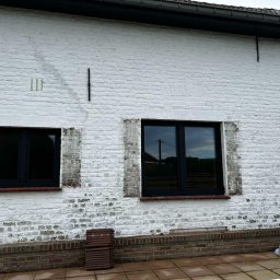 MODERN THERMO BUILDINGS SPÓŁKA Z OGRANICZONĄ ODPOWIEDZIALNOŚCIĄ - Częściowy widok białej, ceglanej ściany budynku z trzema nowo zamontowanymi, czarnymi oknami. Widoczne ślady po poprzednich oknach oraz stos płytek ceramicznych przy ścianie.