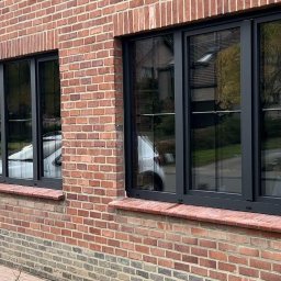 MODERN THERMO BUILDINGS SPÓŁKA Z OGRANICZONĄ ODPOWIEDZIALNOŚCIĄ - Dwa czarne okna z trzema szybami zamontowane w ścianie z czerwonej cegły, w szybach odbija się biały samochód i budynki.