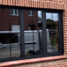 MODERN THERMO BUILDINGS SPÓŁKA Z OGRANICZONĄ ODPOWIEDZIALNOŚCIĄ - Nowe, trzyskrzydłowe okno z czarną ramą zamontowane w ścianie z czerwonej cegły, odbijające w szybie biały samochód dostawczy i fragment budynku.