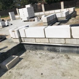 MODERN THERMO BUILDINGS SPÓŁKA Z OGRANICZONĄ ODPOWIEDZIALNOŚCIĄ - Wznoszenie ścian fundamentowych z bloczków betonowych na przygotowanym terenie budowy domu jednorodzinnego, widoczne ułożone warstwy bloczków i zapas materiału budowlanego.