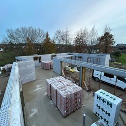 MODERN THERMO BUILDINGS SPÓŁKA Z OGRANICZONĄ ODPOWIEDZIALNOŚCIĄ - Wznoszenie ścian parteru domu z bloczków betonu komórkowego H+H w systemie isoblock, widoczne palety z bloczkami, ściany w trakcie budowy, widok z góry na plac budowy w pochmurny dzień.