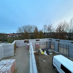 MODERN THERMO BUILDINGS SPÓŁKA Z OGRANICZONĄ ODPOWIEDZIALNOŚCIĄ - Wznoszenie ścian domu z szarych bloczków isoblock na wylewce fundamentowej, widoczne okna zbrojone, palety z materiałami budowlanymi i biały samochód dostawczy na placu budowy.