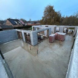 MODERN THERMO BUILDINGS SPÓŁKA Z OGRANICZONĄ ODPOWIEDZIALNOŚCIĄ - Budowa domu z wykorzystaniem szalunku isoblock, widoczne ściany wzniesione do wysokości okien, zbrojenia, oraz palety z cegłami, w tle zabudowa jednorodzinna i drzewa.