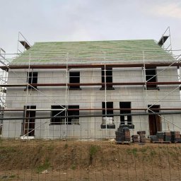 MODERN THERMO BUILDINGS SPÓŁKA Z OGRANICZONĄ ODPOWIEDZIALNOŚCIĄ - Budowa domu z pustaków isoblock, widoczne rusztowania i przygotowane dachówki, teren ogrodzony siatką.