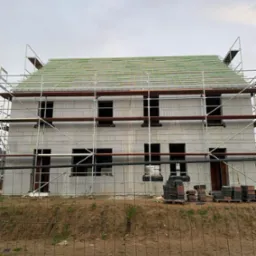 Budowa domu z pustaków isoblock, widoczne rusztowania i przygotowane dachówki, teren ogrodzony siatką.