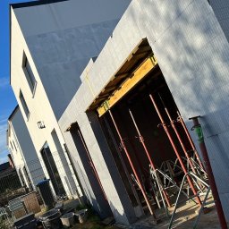 MODERN THERMO BUILDINGS SPÓŁKA Z OGRANICZONĄ ODPOWIEDZIALNOŚCIĄ - Budowa domu w systemie isoblock: widok na fragment ściany z widocznymi podporami i szalunkiem, z fragmentem elewacji w tle, pod błękitnym niebem.