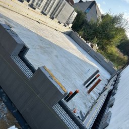 MODERN THERMO BUILDINGS SPÓŁKA Z OGRANICZONĄ ODPOWIEDZIALNOŚCIĄ - Budowa fundamentów domu z wykorzystaniem szalunków systemowych Isoblock, widoczne rury instalacyjne i częściowo wylana płyta fundamentowa, w tle budynek mieszkalny i zieleń.