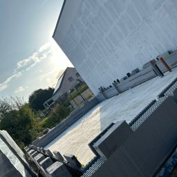 MODERN THERMO BUILDINGS SPÓŁKA Z OGRANICZONĄ ODPOWIEDZIALNOŚCIĄ - Budowa domu w systemie isoblock: widok na fundamenty i ściany wznoszone z szarych, modułowych elementów, z widocznym zarysem sąsiedniego domu w tle.