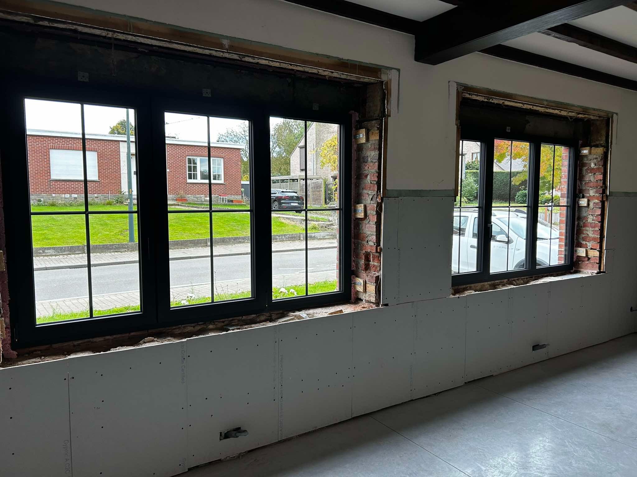 Wnętrze w trakcie remontu z dwoma oknami z czarnymi ramami, widoczne surowe ściany z cegły i płyty gipsowo-kartonowej, na zewnątrz budynki mieszkalne i samochody