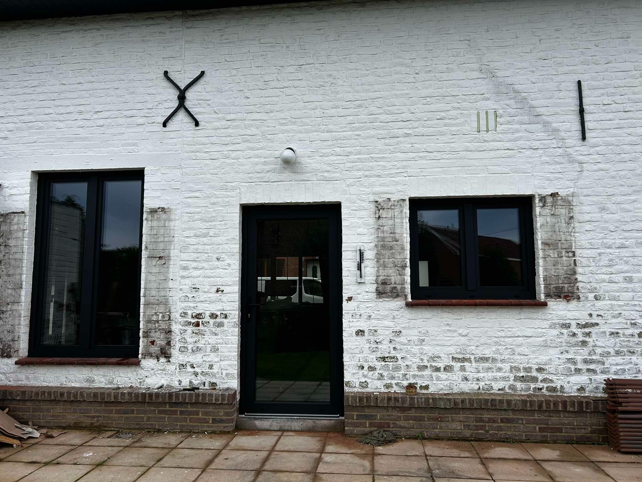 Biała, ceglana ściana budynku z zamontowanymi czarnymi oknami PCV i drzwiami. Na ścianie widoczne dekoracje w postaci metalowych elementów.