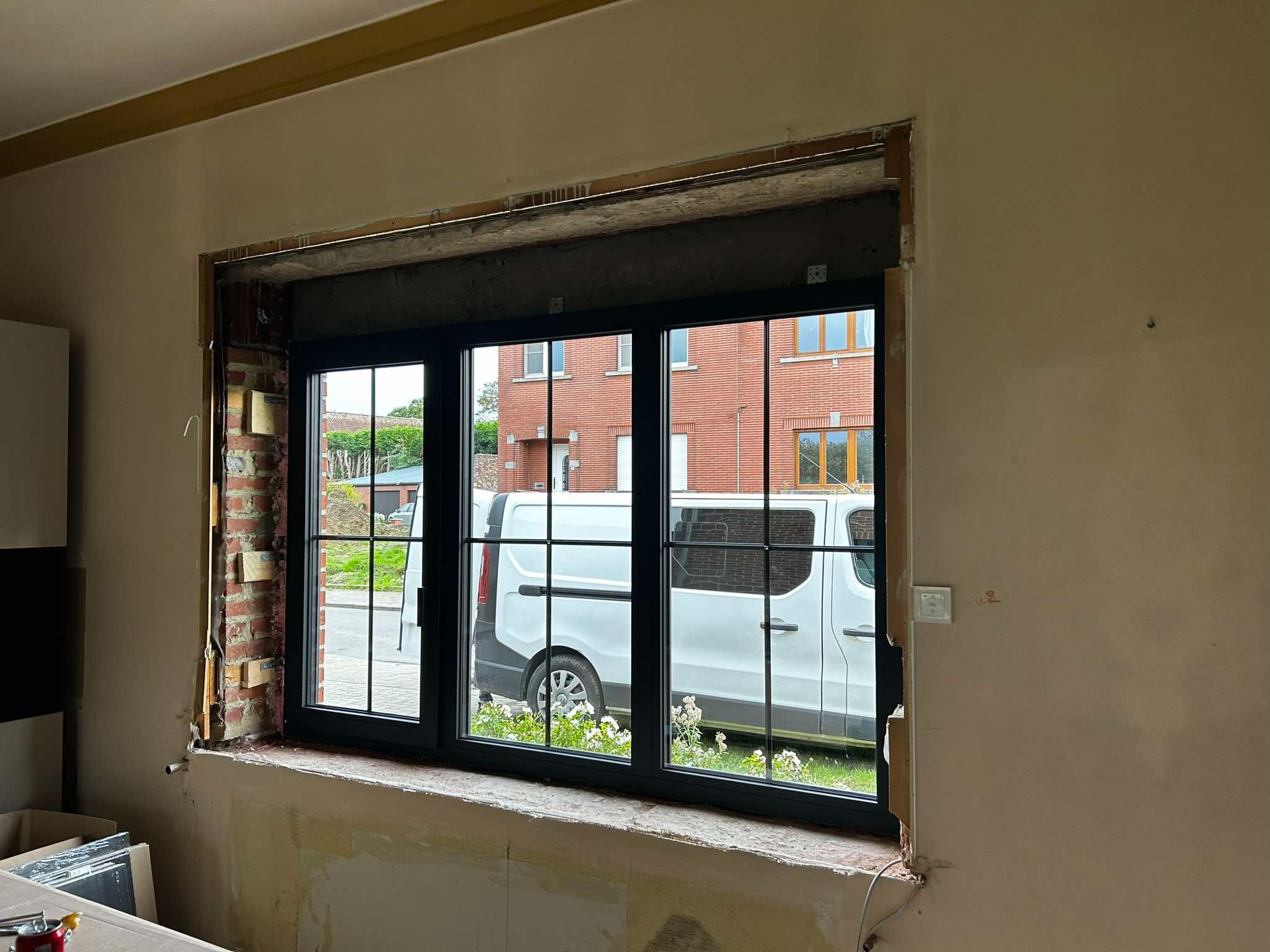 Montaż czarnej stolarki okiennej z kratownicą w domu w Chruślinie, widok na ceglany budynek przez okno, biały samochód dostawczy zaparkowany na zewnątrz.