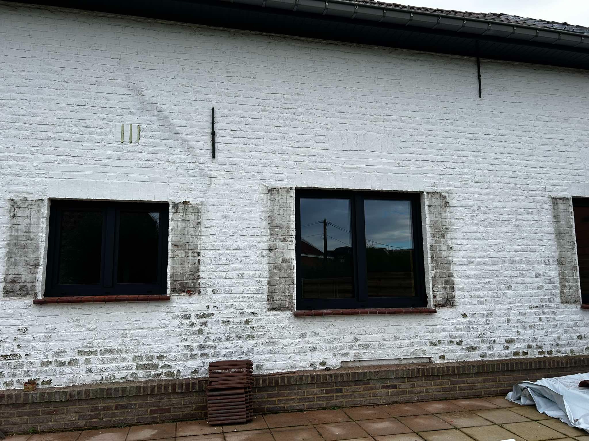 Częściowy widok białej, ceglanej ściany budynku z trzema nowo zamontowanymi, czarnymi oknami. Widoczne ślady po poprzednich oknach oraz stos płytek ceramicznych przy ścianie.