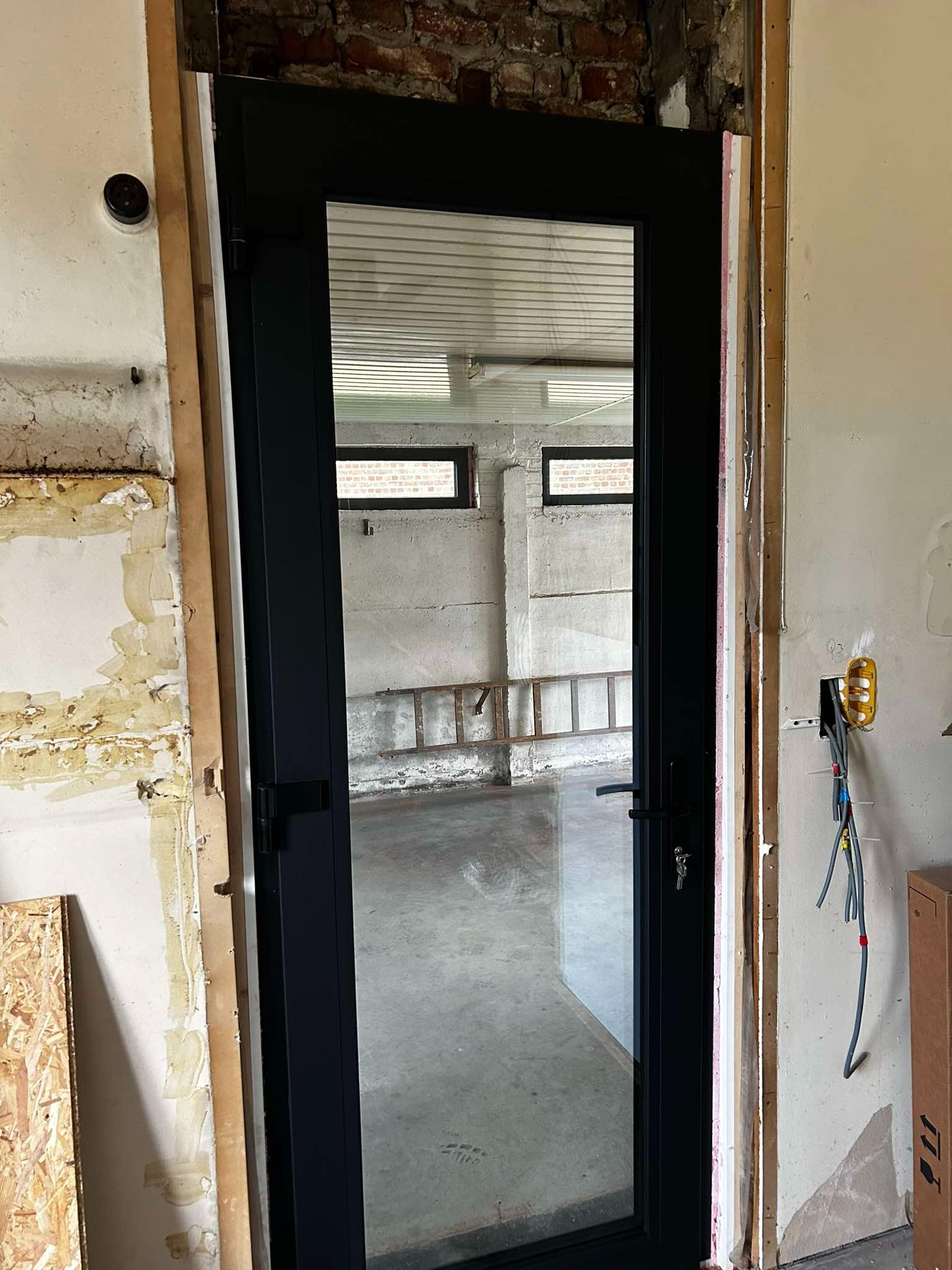 Czarne drzwi balkonowe z szybą zamontowane w surowym otworze, widoczne ślady po poprzedniej instalacji, przewody elektryczne wystające ze ściany, w tle drabina i okna piwniczne.