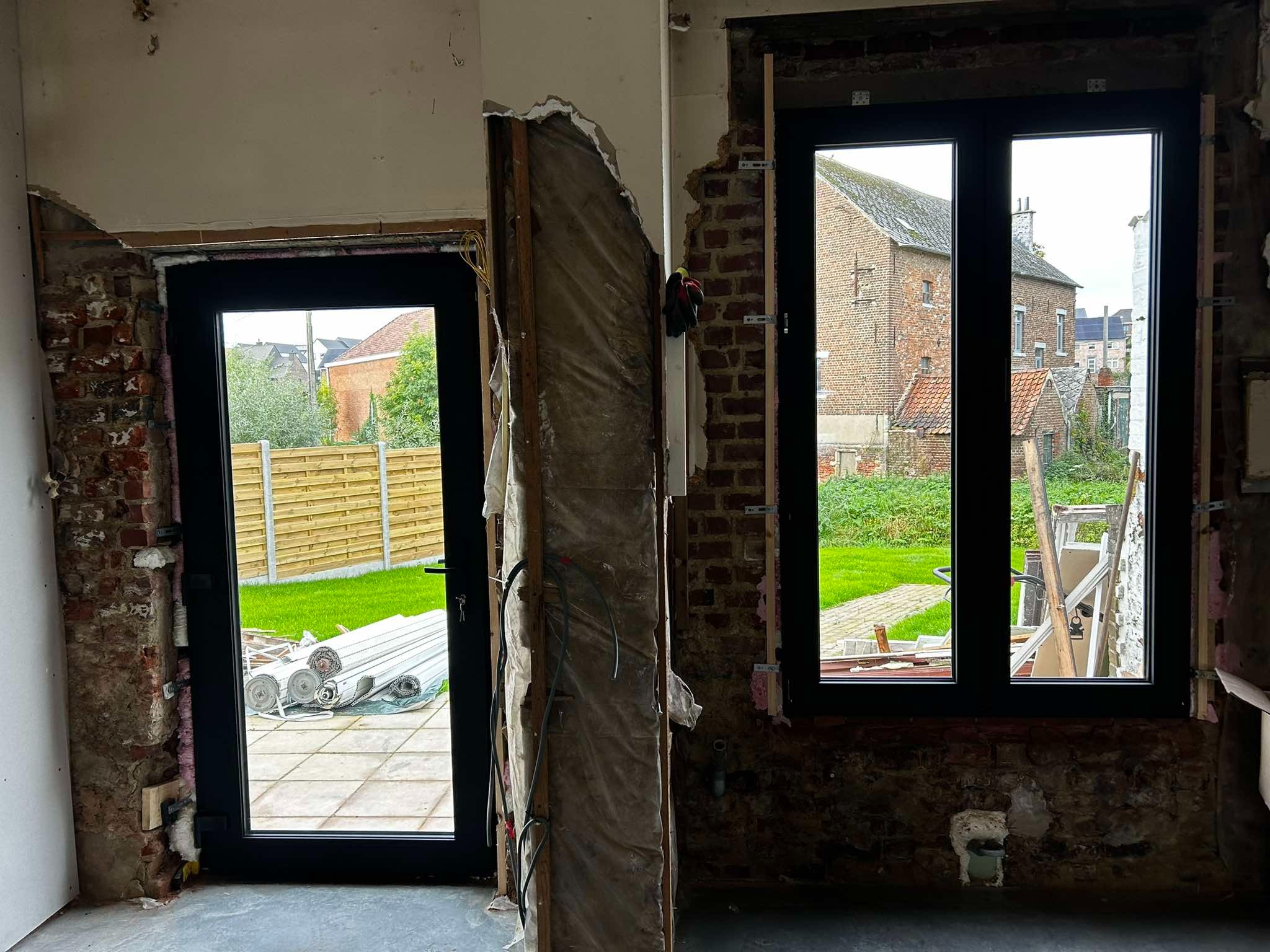 Wnętrze w trakcie remontu z zamontowaną czarną stolarką okienną i drzwiową, widoczne odsłonięte cegły, instalacje i materiały budowlane na zewnątrz.