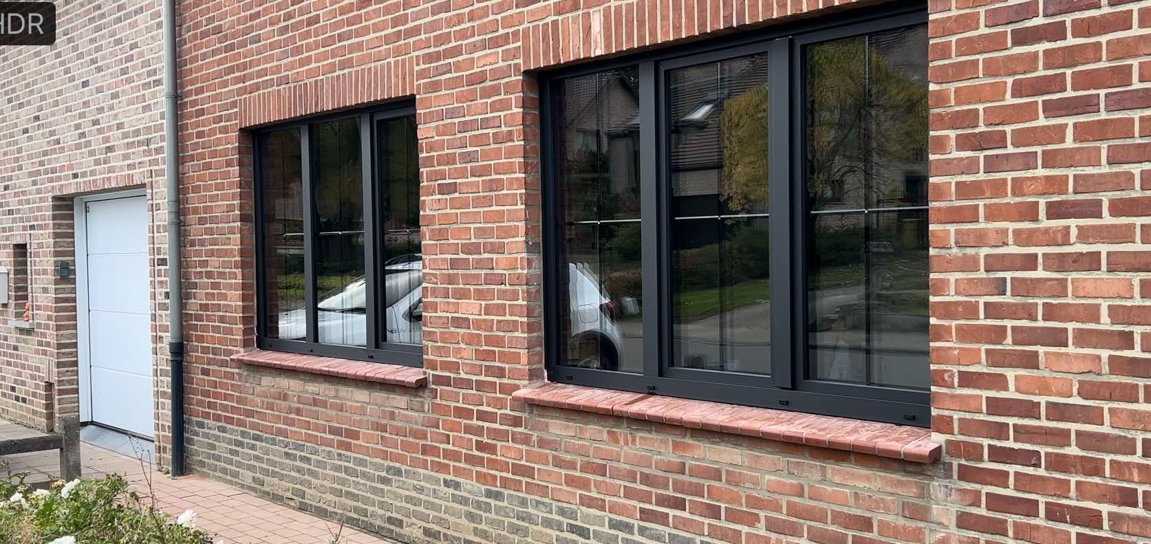 Dwa czarne okna z trzema szybami zamontowane w ścianie z czerwonej cegły, w szybach odbija się biały samochód i budynki.