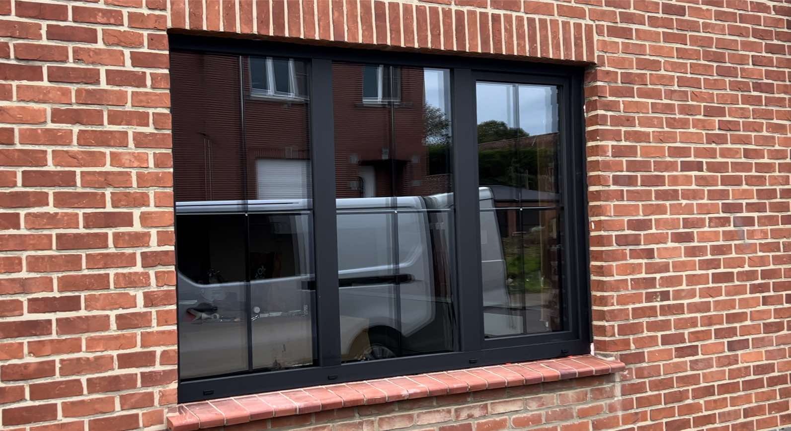 Nowe, trzyskrzydłowe okno z czarną ramą zamontowane w ścianie z czerwonej cegły, odbijające w szybie biały samochód dostawczy i fragment budynku.