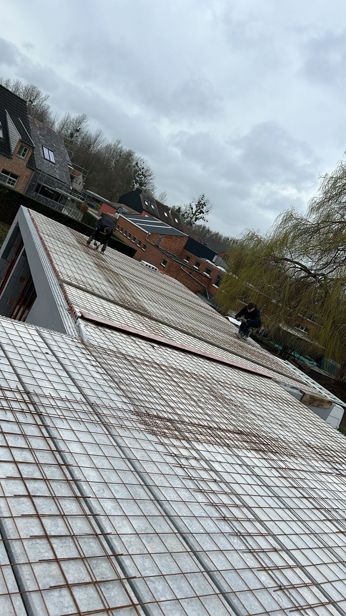 Dwóch pracowników montuje zbrojenie na płycie fundamentowej domu modułowego, widok z góry, w tle zabudowa jednorodzinna i pochmurne niebo.