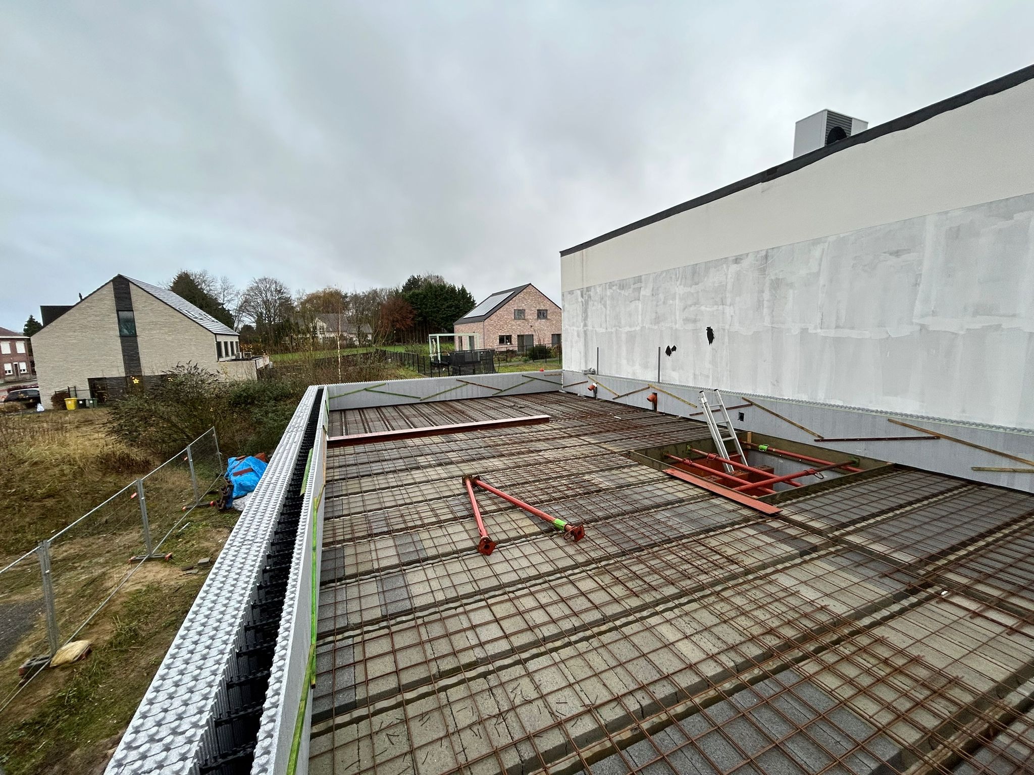 Zbrojenie stropu przygotowane do zalania betonem, widoczne szalunki z bloczków isoblock, podpory i drabina, w tle domy jednorodzinne i pochmurne niebo.