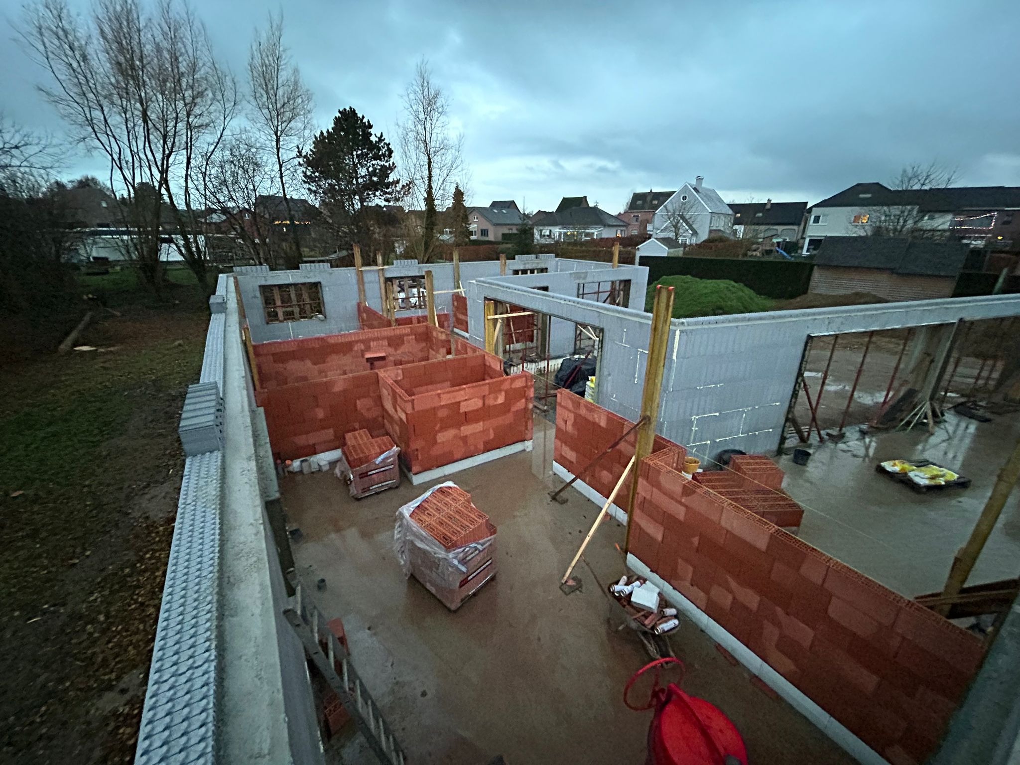 Wznoszenie ścian domu z czerwonej cegły modułowej w systemie isoblock, widok z góry na plac budowy w pochmurny dzień.