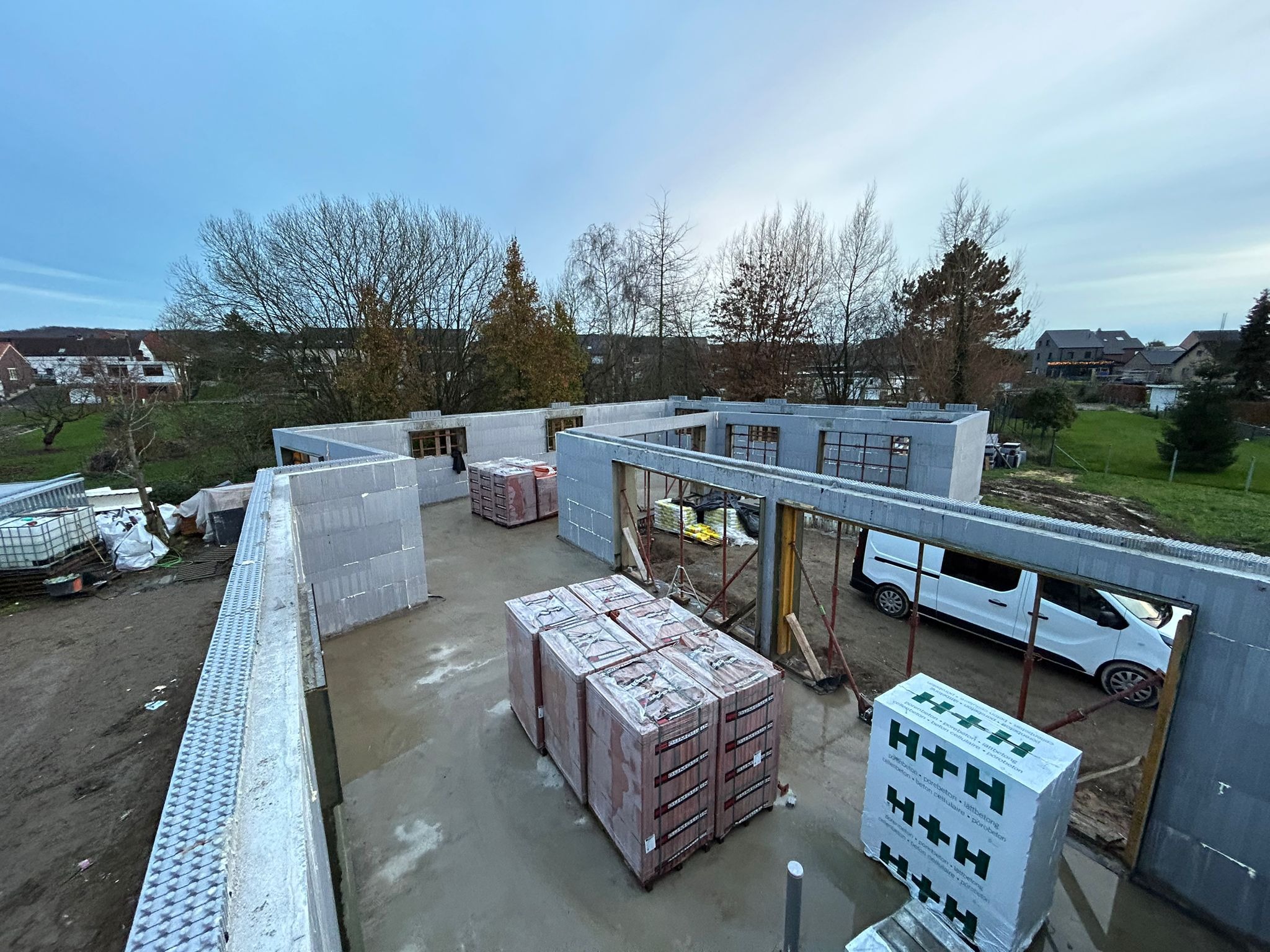 Wznoszenie ścian parteru domu z bloczków betonu komórkowego H+H w systemie isoblock, widoczne palety z bloczkami, ściany w trakcie budowy, widok z góry na plac budowy w pochmurny dzień.