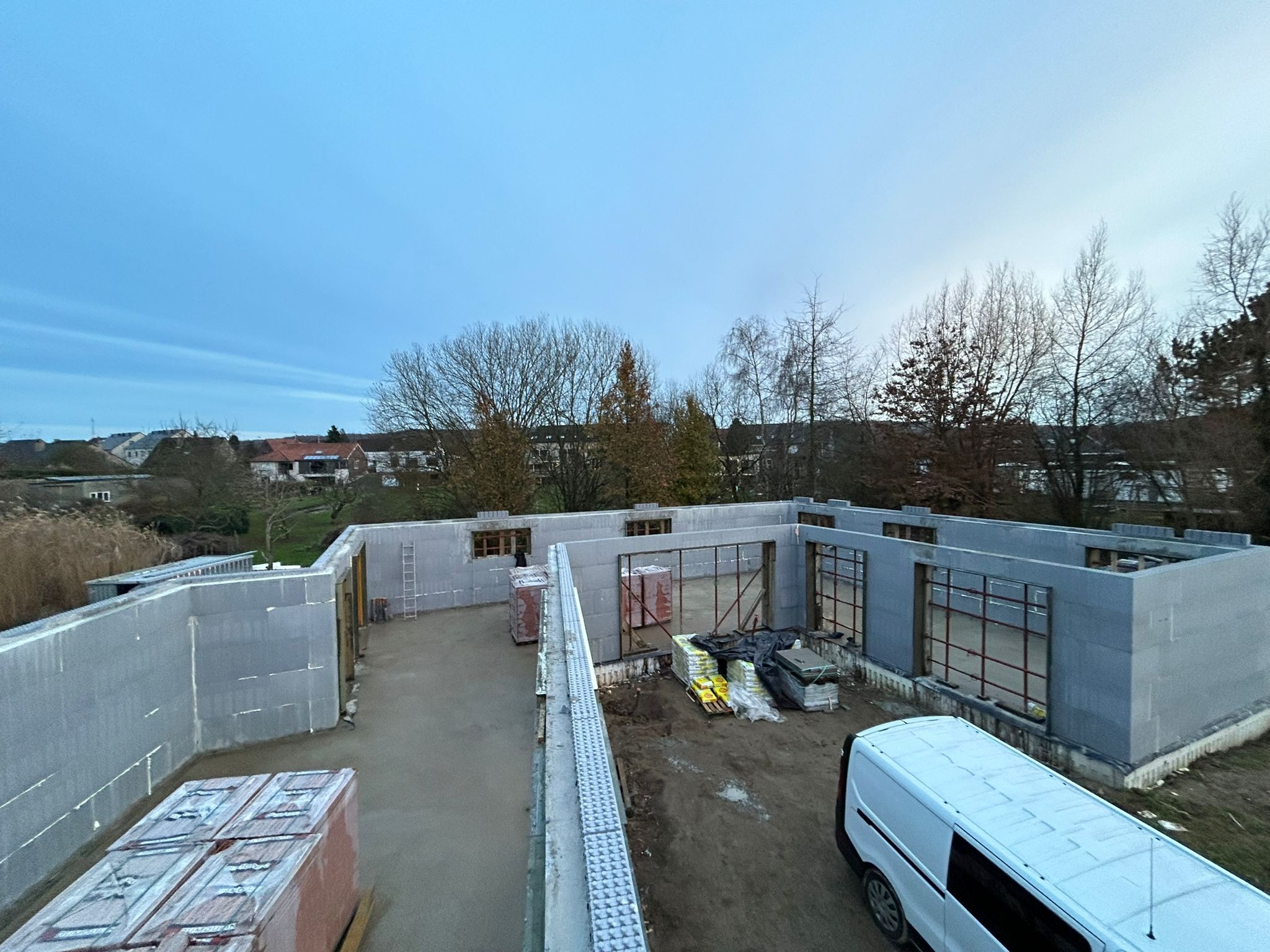 Wznoszenie ścian domu z szarych bloczków isoblock na wylewce fundamentowej, widoczne okna zbrojone, palety z materiałami budowlanymi i biały samochód dostawczy na placu budowy.