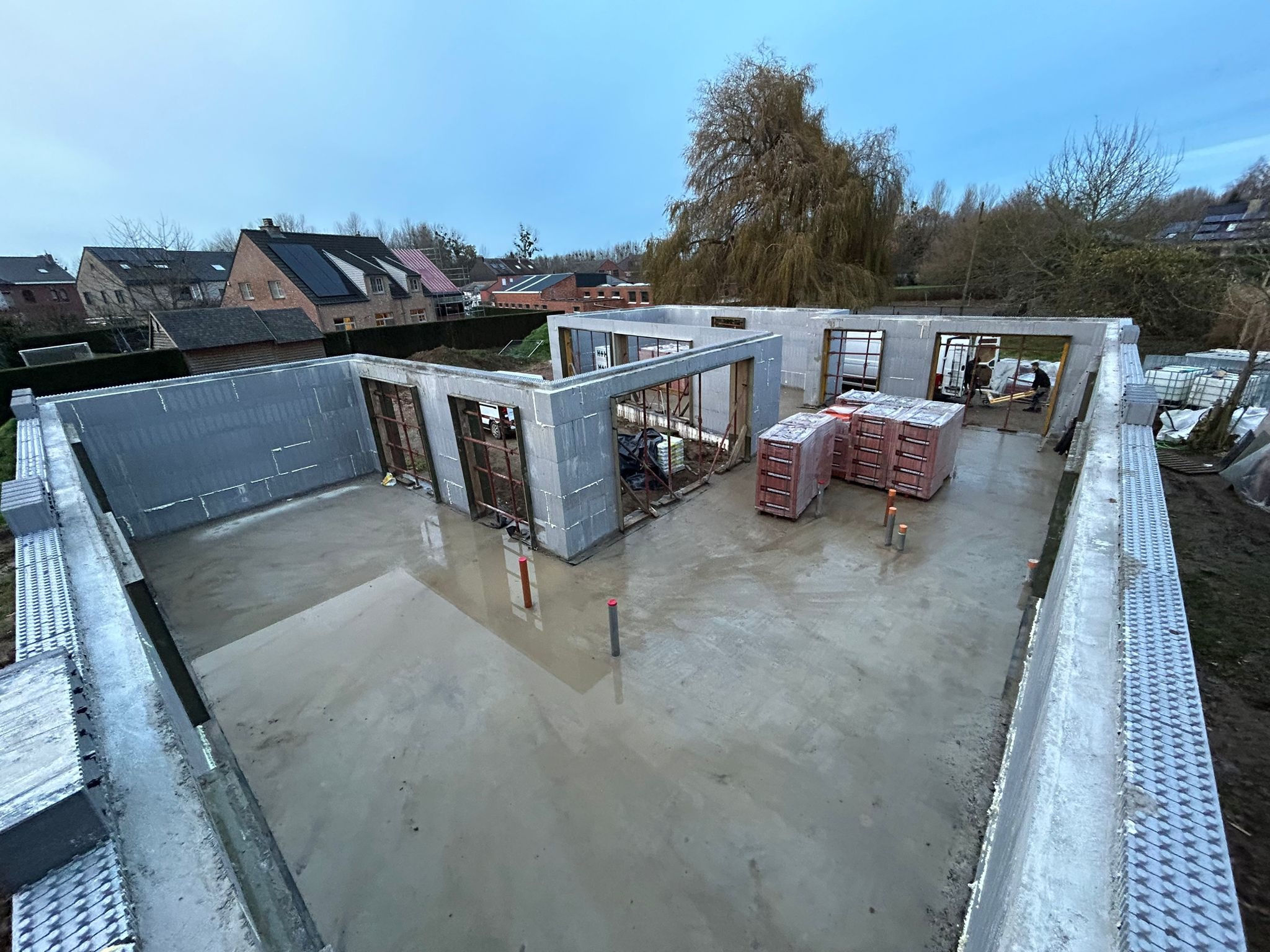 Budowa domu z wykorzystaniem szalunku isoblock, widoczne ściany wzniesione do wysokości okien, zbrojenia, oraz palety z cegłami, w tle zabudowa jednorodzinna i drzewa.