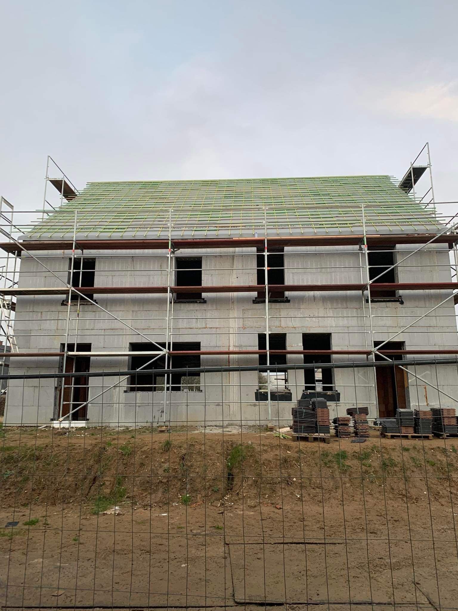 Budowa domu z pustaków isoblock, widoczne rusztowania i przygotowane dachówki, teren ogrodzony siatką.