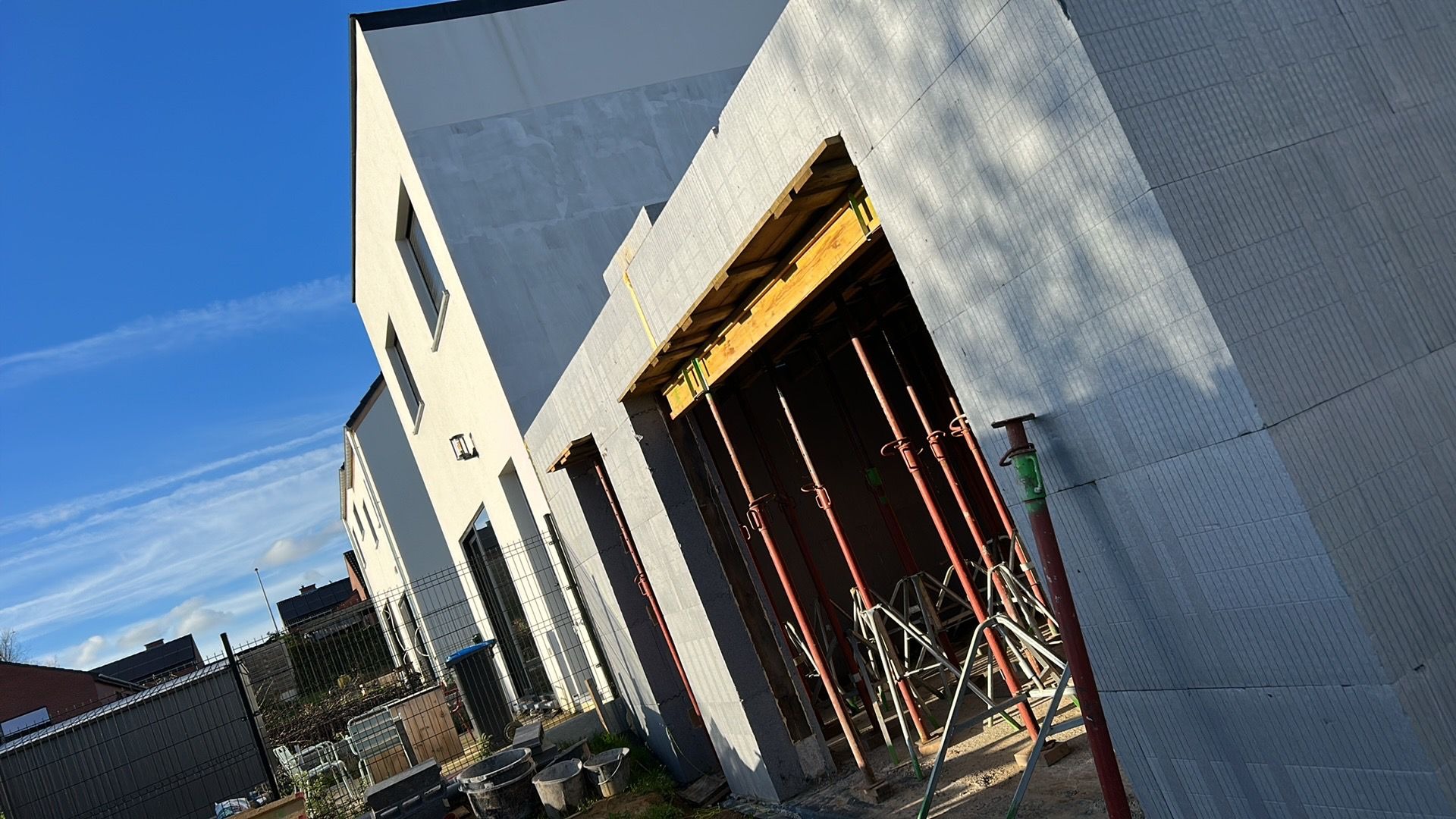 Budowa domu w systemie isoblock: widok na fragment ściany z widocznymi podporami i szalunkiem, z fragmentem elewacji w tle, pod błękitnym niebem.