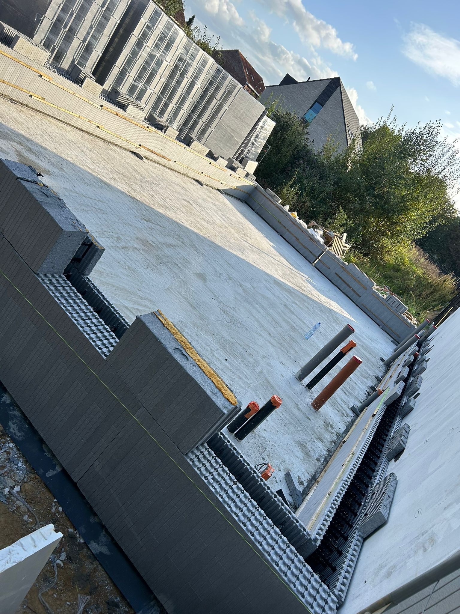 Budowa fundamentów domu z wykorzystaniem szalunków systemowych Isoblock, widoczne rury instalacyjne i częściowo wylana płyta fundamentowa, w tle budynek mieszkalny i zieleń.