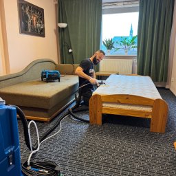 A.P. Alfa Sp. z o.o. - Uśmiechnięty mężczyzna w czarnej koszulce, klęczący na wzorzystym dywanie, czyści materac na drewnianym łóżku przy użyciu profesjonalnego sprzętu do ekstrakcyjnego czyszczenia; w tle widoczna...