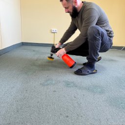 A.P. Alfa Sp. z o.o. - Mężczyzna w klapkach i skarpetkach, w pozycji kucznej, czyści jasnoniebieski dywan przy użyciu profesjonalnego urządzenia z pomarańczowym zbiornikiem na płyn i okrągłą szczotką. Widoczne ciemne...