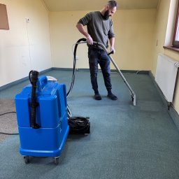 A.P. Alfa Sp. z o.o. - Mężczyzna w rękawicach czyści zielony dywan w pustym pokoju za pomocą niebieskiego odkurzacza piorącego.