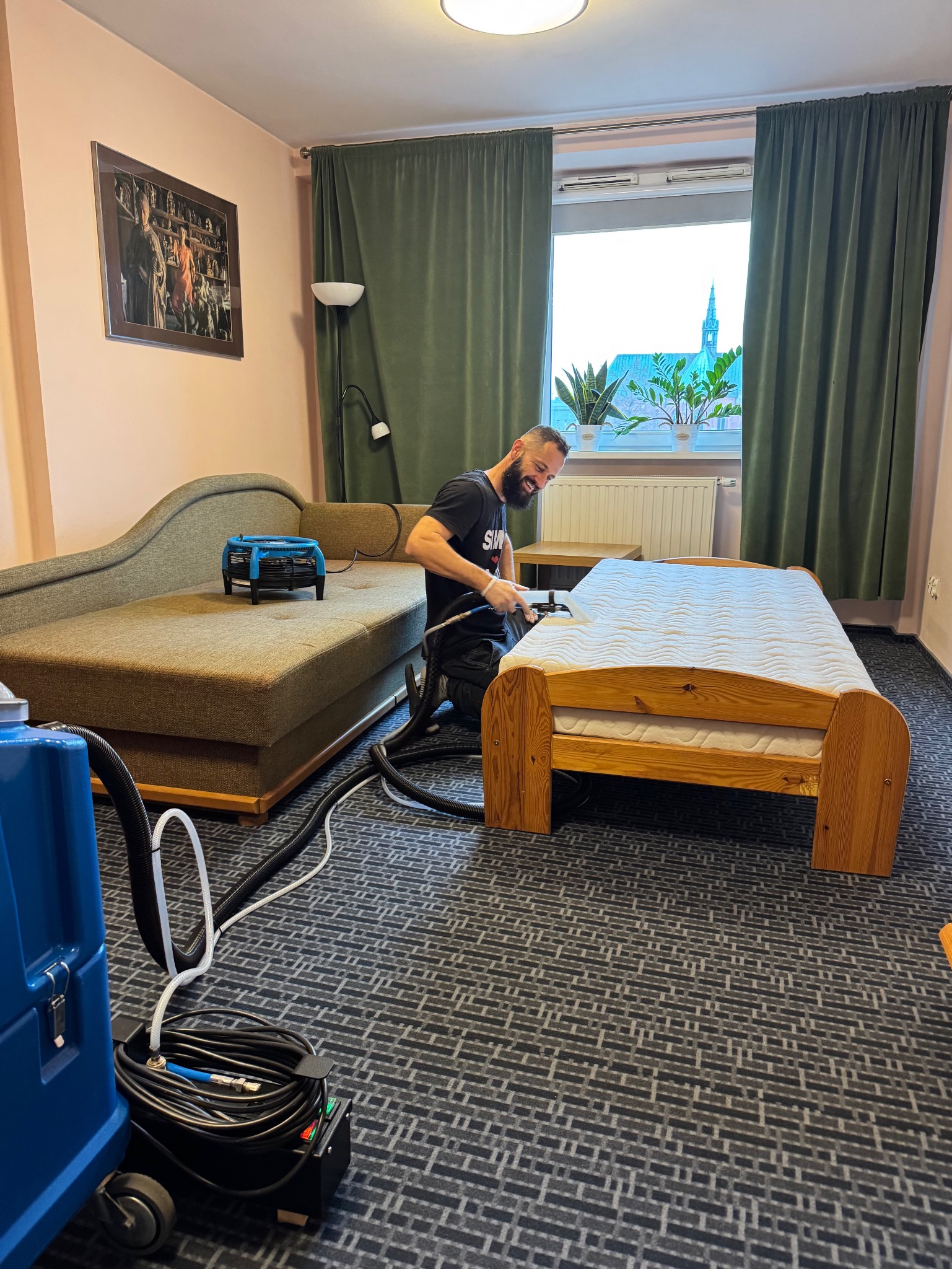 Uśmiechnięty mężczyzna w czarnej koszulce, klęczący na wzorzystym dywanie, czyści materac na drewnianym łóżku przy użyciu profesjonalnego sprzętu do ekstrakcyjnego czyszczenia; w tle widoczna...