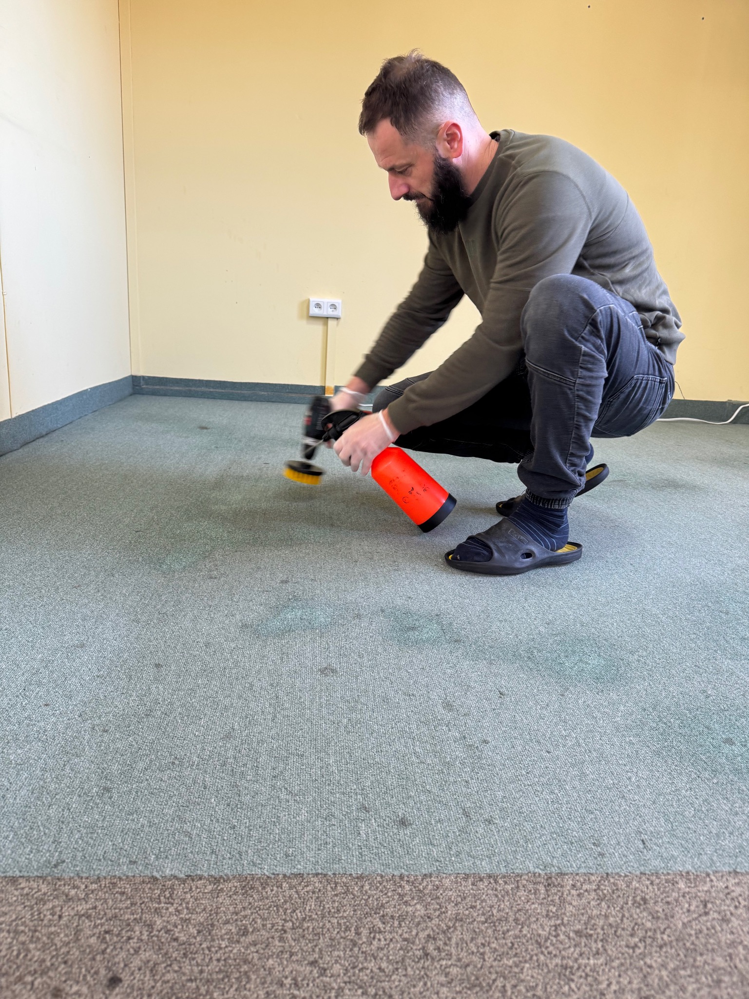 Mężczyzna w klapkach i skarpetkach, w pozycji kucznej, czyści jasnoniebieski dywan przy użyciu profesjonalnego urządzenia z pomarańczowym zbiornikiem na płyn i okrągłą szczotką. Widoczne ciemne...