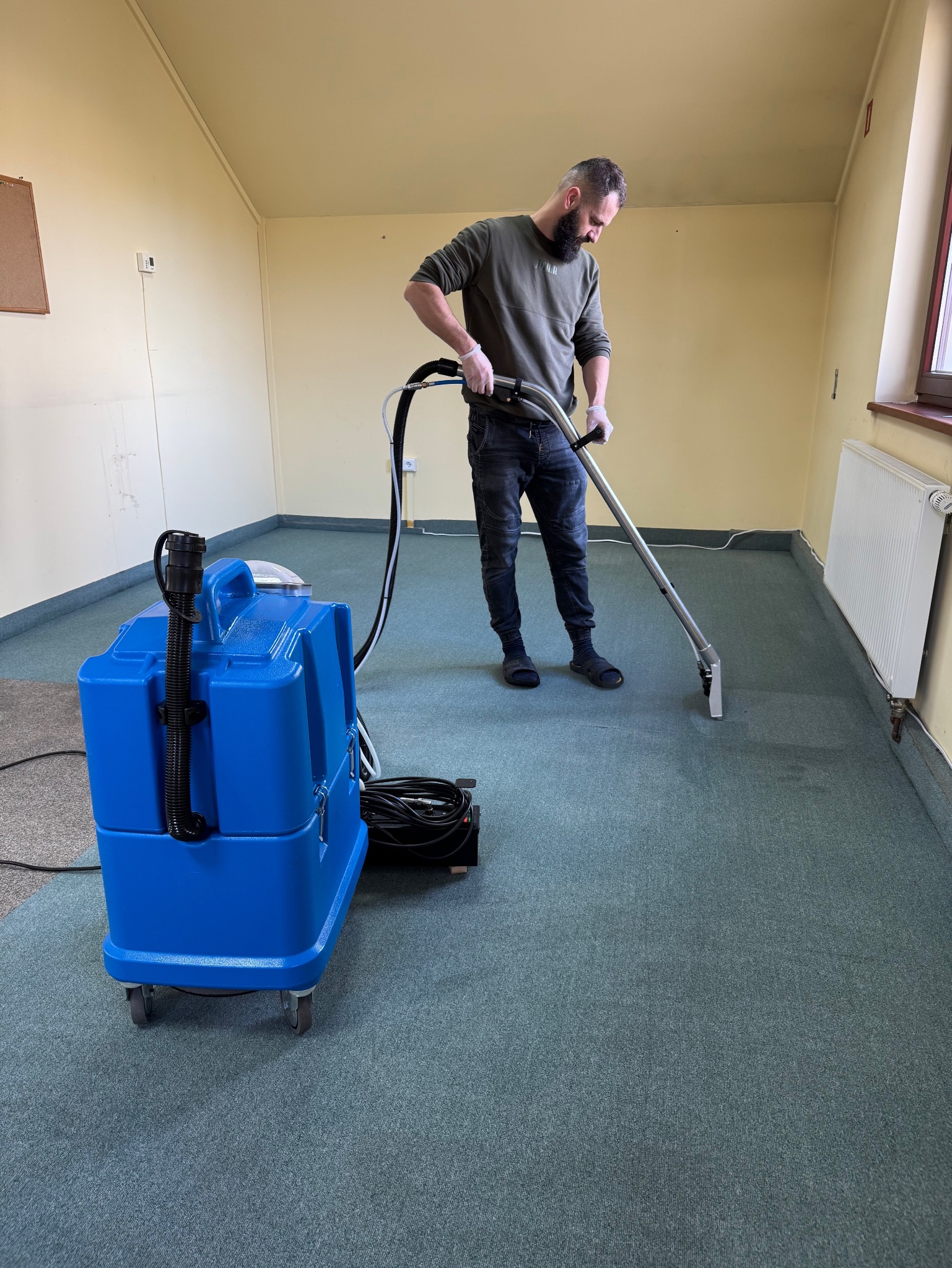 Mężczyzna w rękawicach czyści zielony dywan w pustym pokoju za pomocą niebieskiego odkurzacza piorącego.