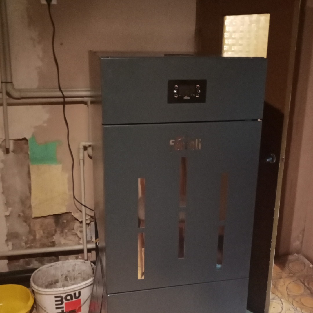 Szary piec na paliwo stałe marki Ferroli w kotłowni z widocznymi fragmentami instalacji grzewczej i wiaderkiem po farbie.