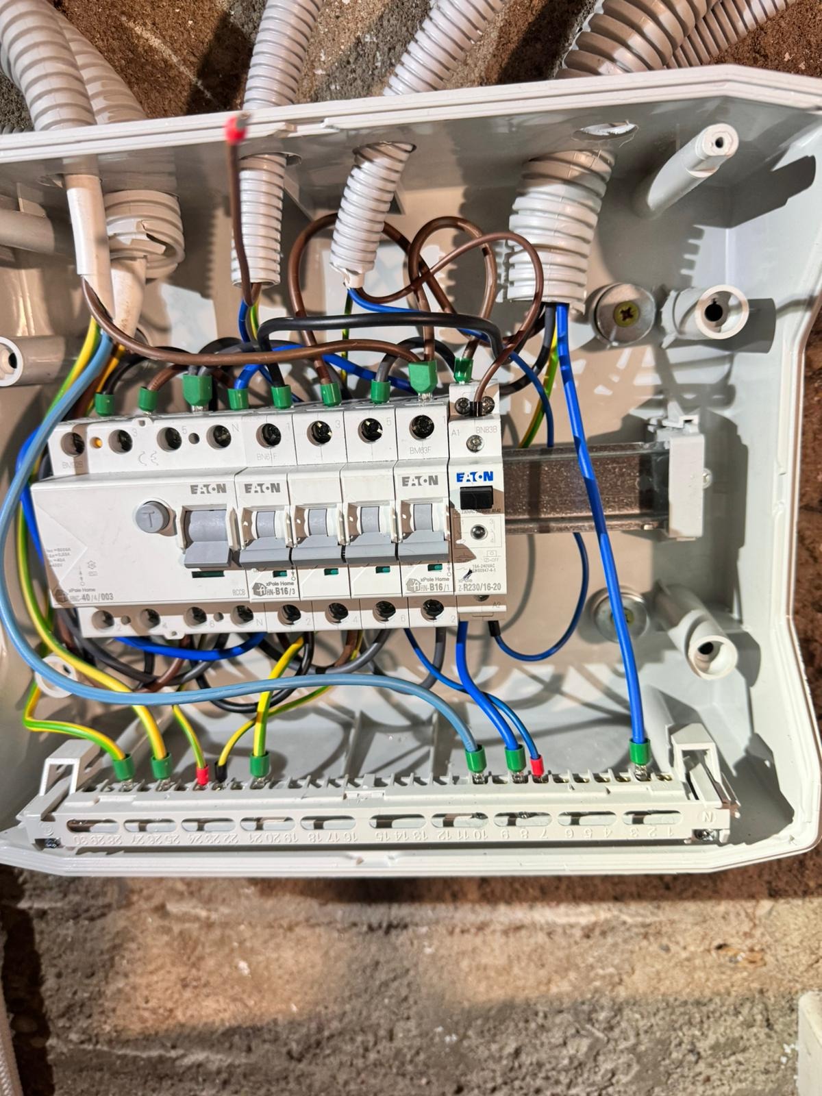 Otwarta skrzynka elektryczna z zamontowanymi bezpiecznikami marki Eaton, widoczne liczne przewody w różnych kolorach (niebieski, żółto-zielony, brązowy, czarny) podłączone do listwy zaciskowej...